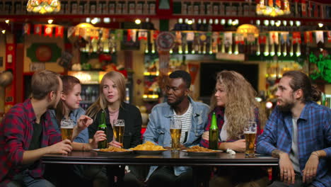 Eine-Multiethnische-Gruppe-Junger-Männer-Und-Frauen-Trinkt-In-Einer-Bar-Bier,-Isst-Chips-Und-Diskutiert-Fröhlich-über-Die-Universität.-Sie-Lachen-über-Einen-Witz.